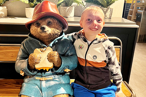 A young blonde haired boy is sat on an indoor bench next to a statue of Paddington Bear eating a marmalade sandwich and wearing his traditional blue dufflecoat and red hat. The smiling boy has a port wine birthmark on the left side of his face. He is wearing a brown and white hoodie with camouflage sleeves and blue trousers. nThere are a number of plants on a unit behind the bench.