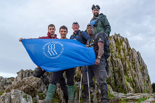 There are five men standing on the rocky peak of a mountain in Wales. They are all wearing sturdy climbing boots and clothing. Three of them have headlamps on their foreheads, one has a set of walking sticks and they are collectively holding a blue flag with the Sturge Weber Uk logo on it. The sky is overcast and it looks a bit windy.