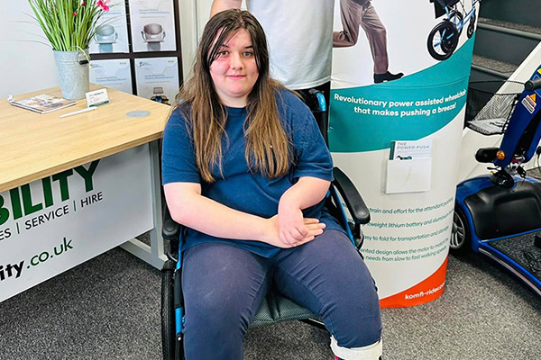 A young lady with long brown hair wearing a blue top and blue trousers is sat in a new wheelchair. There is a table to her left with a plant on it, a man is tanding behind her with a promotional stand and motorised wheelchair to her right.