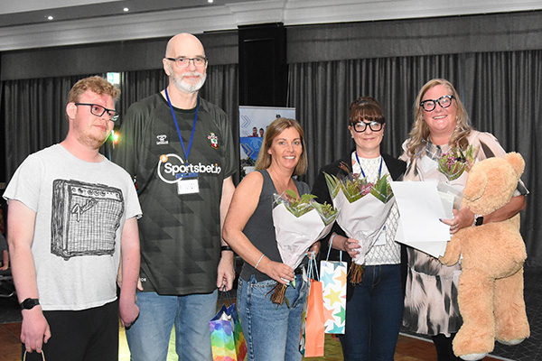 This shows five people, two men and three women who make up part of the Sturge Weber UK Trustees and Volunteers. The women are each holding a bunch of flowers and one has a large teddy bear under her arm. One of the men has aport wine birthmark on his face.