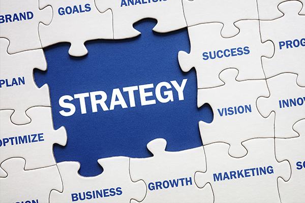 This image shows 4 missong pieces of a white jigsaw puzzle that have many words on each of the other pieces, such as business, growth, marketing, vision, success. Each piece has one word on it. The section with the four missing pieces has a blue background with the word Strategy in bold white capitals printed on.