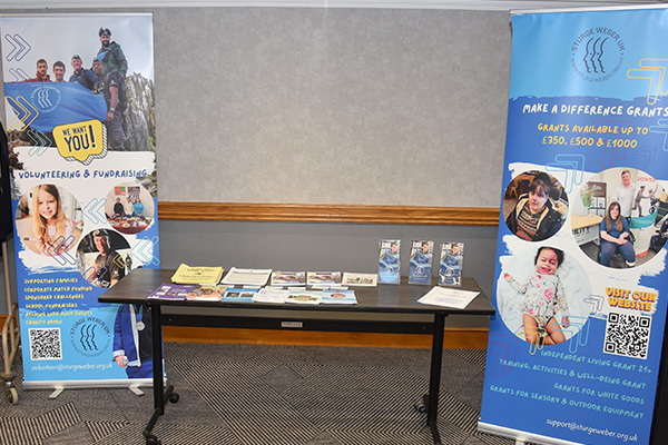 The picture shows a two promotional banners encouraging volunteers to help the charity either side of a wooden and metal table with leaflets and booklets about the charity displayed on it. The table and banners are stood on a brown carpeted floor.